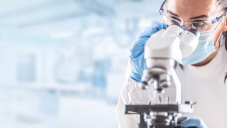 Female lab technician in protective glasses, gloves and face mask sits next to a microscope in laboratory.