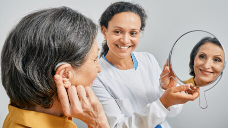 Mature woman with BTE hearing aid looks at himself in mirror held by smiling audiologist, and tries on hearing device. Stylish hearing device