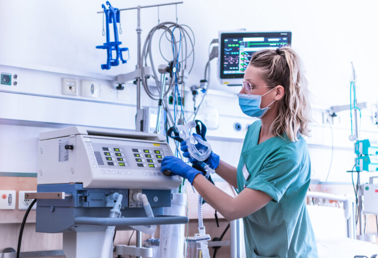 COVID19 / 2019-ncov concept: nurse, wearing a surgical mask, checks the settings of a mechanical ventilation machine, which is seen in the foreground. therapy used for lung breathing, in intensive care.